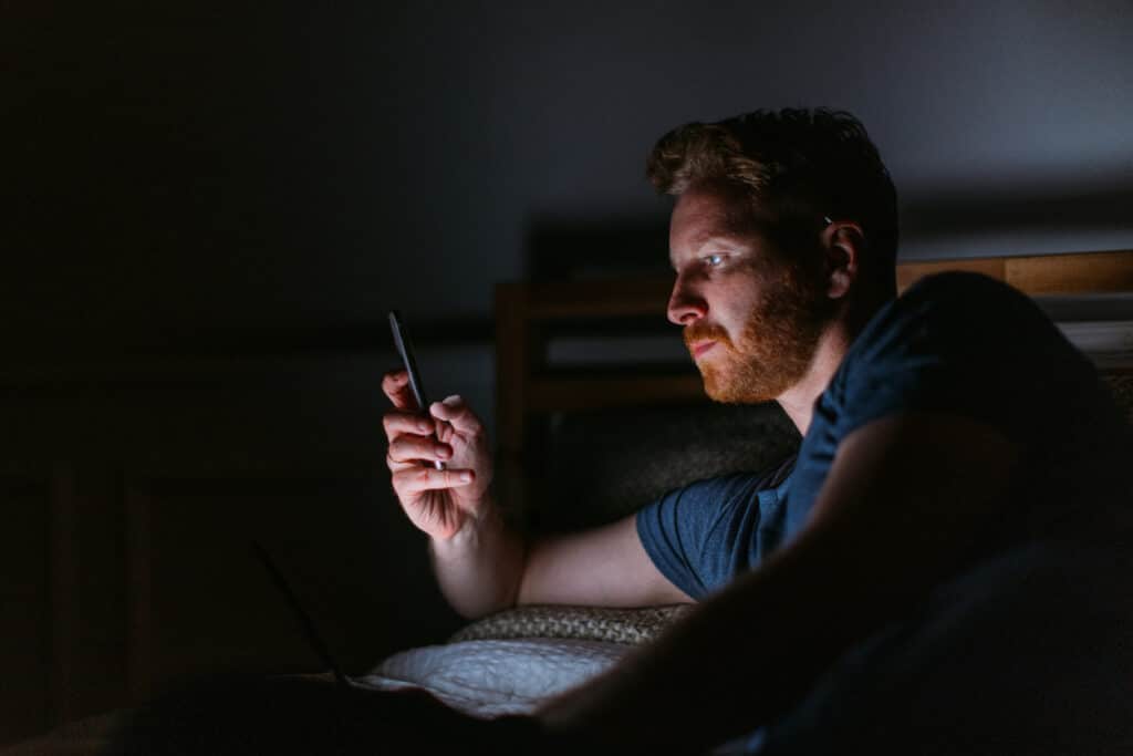 A contemplative Caucasian man using his smartphone while resting in bed at night, grappling with sex addiction.