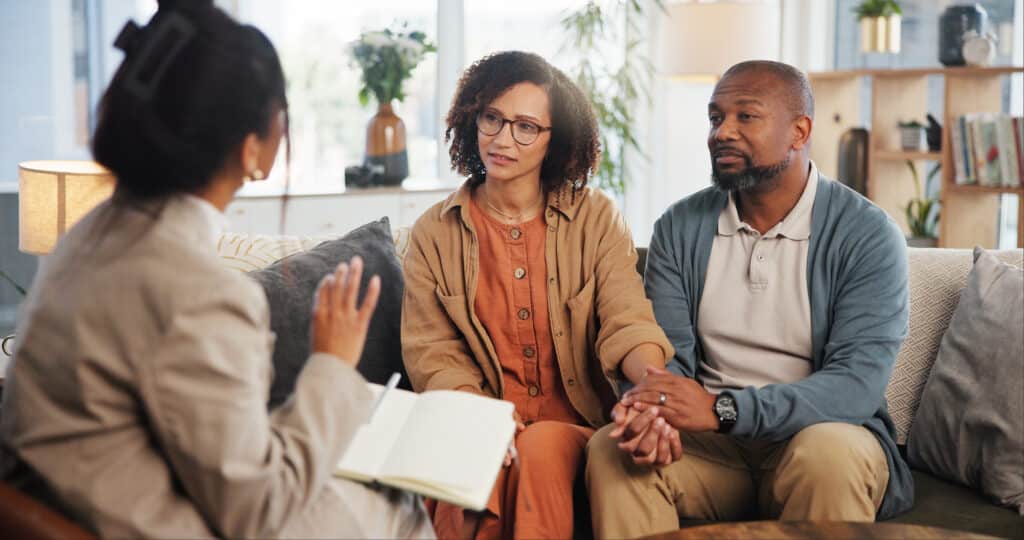 Couple, therapy and holding hands with discussion on sofa for guidance, communication and advice. Consultation, service and therapist with book in office for marriage counseling, support and progress after sex addiction