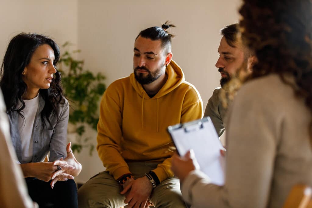 Cut out shot of anxious woman sitting in circle and talking about her mental health struggles with her peers during a group therapy session in rehab.