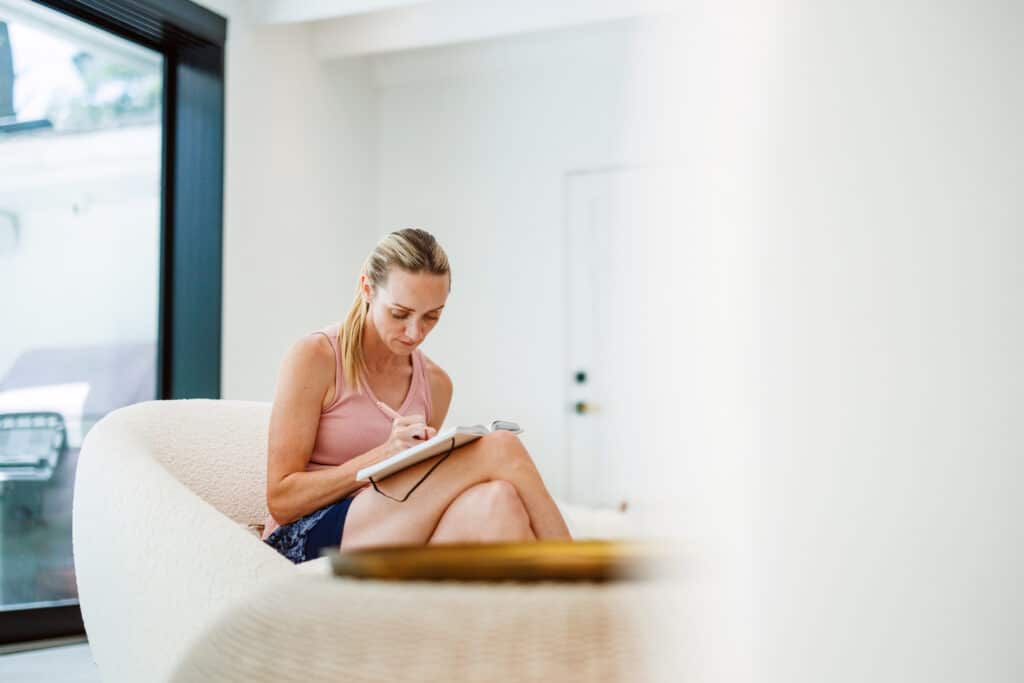 A focused mid-adult Caucasian woman pensively writes in her journal while seated in a relaxed postured chair at a modern rehabilitation center, indicating progress in her recovery journey.