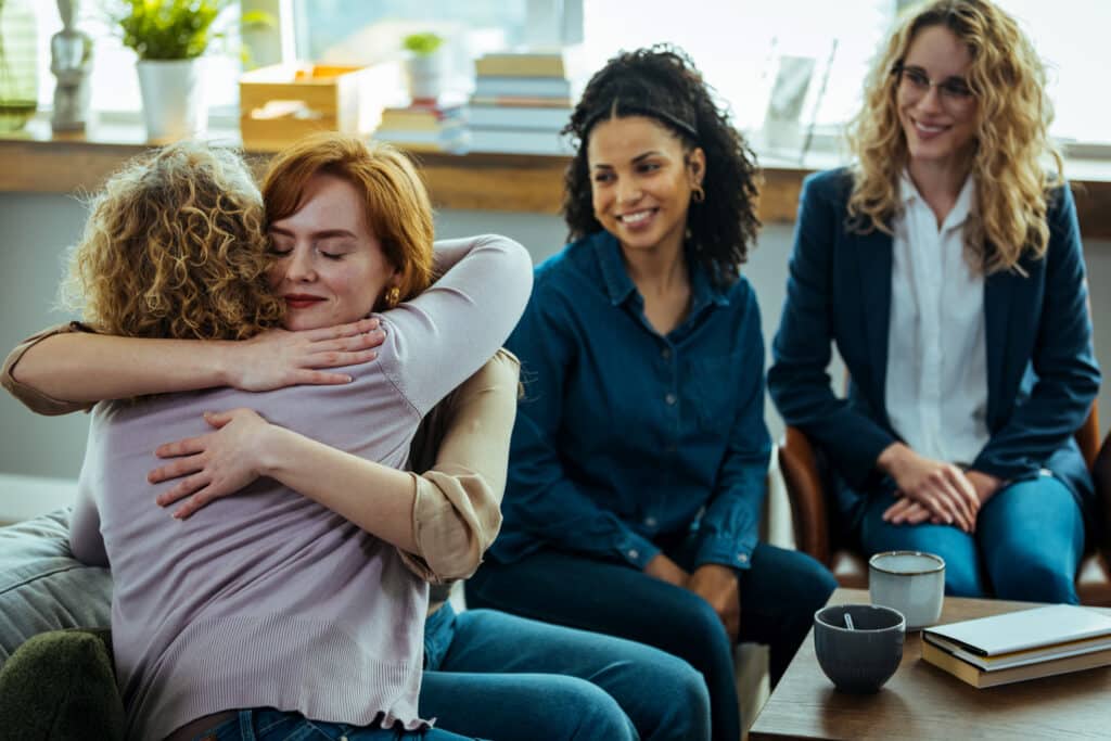 Caring female counselor hugs a female patient during a group therapy session.